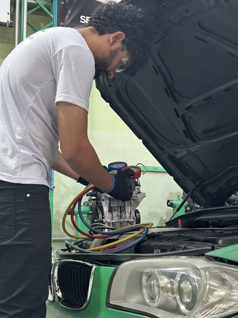 Técnico automotivo realizando manutenção e teste de pressão no sistema de ar-condicionado de um veículo.